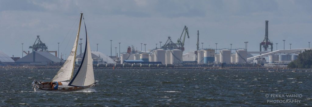 Purjevene merellä, taustalla satama-alue.