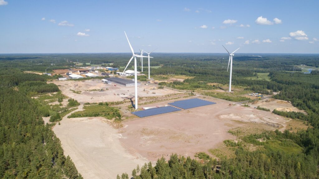 Aurinkopaneeleja ja tuulimyllyjä laajalla hiekkakentällä, jonka ympärillä puustoa.